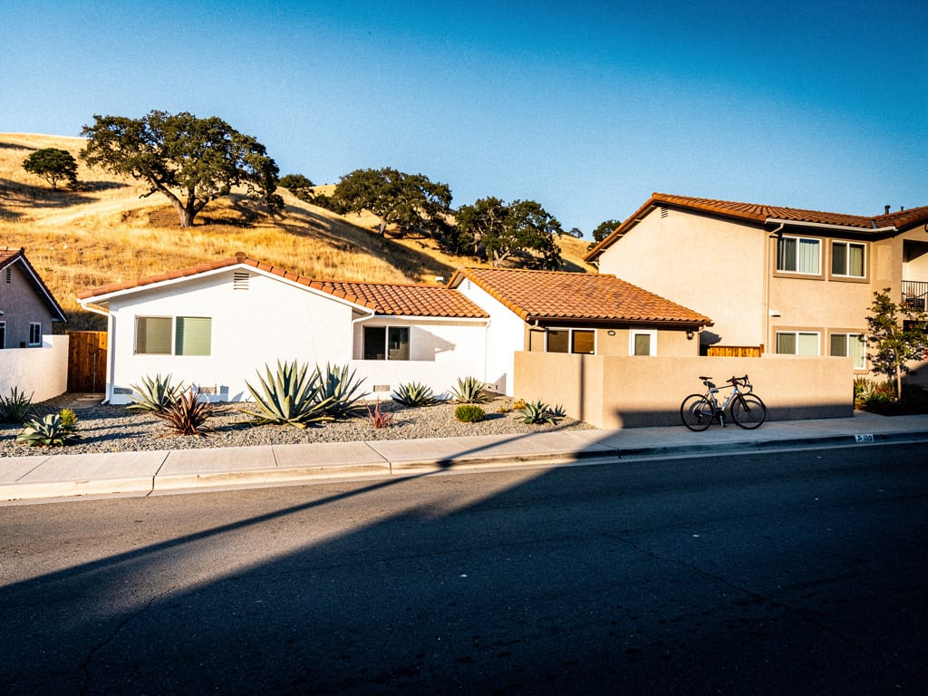 A quiet Central Coast residential street of stucco duplexes and small apartment buildings with golden hills behind